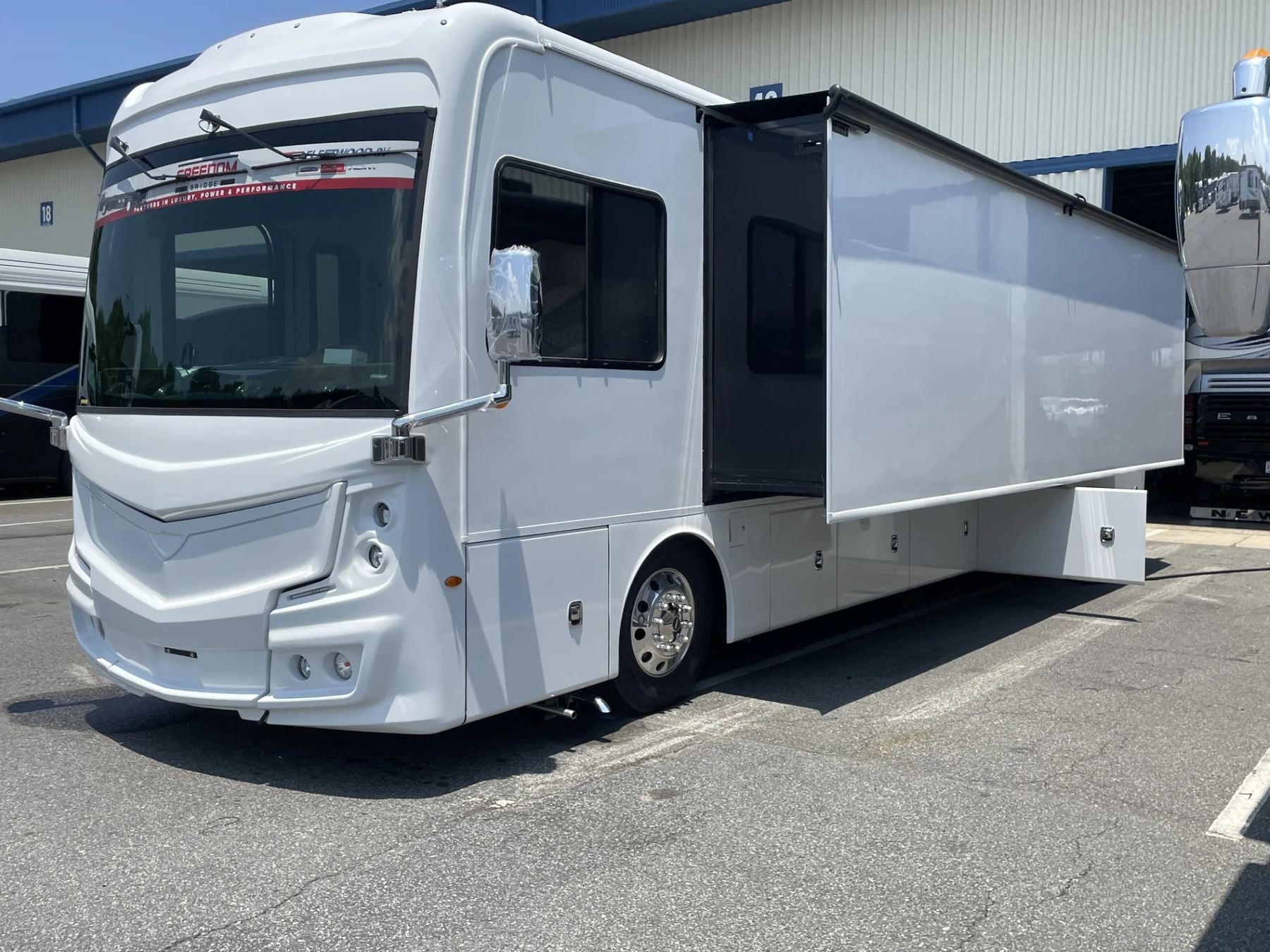 The unwrapped white vehicle parked outside a warehouse, seen from the front corner with its side compartment open.